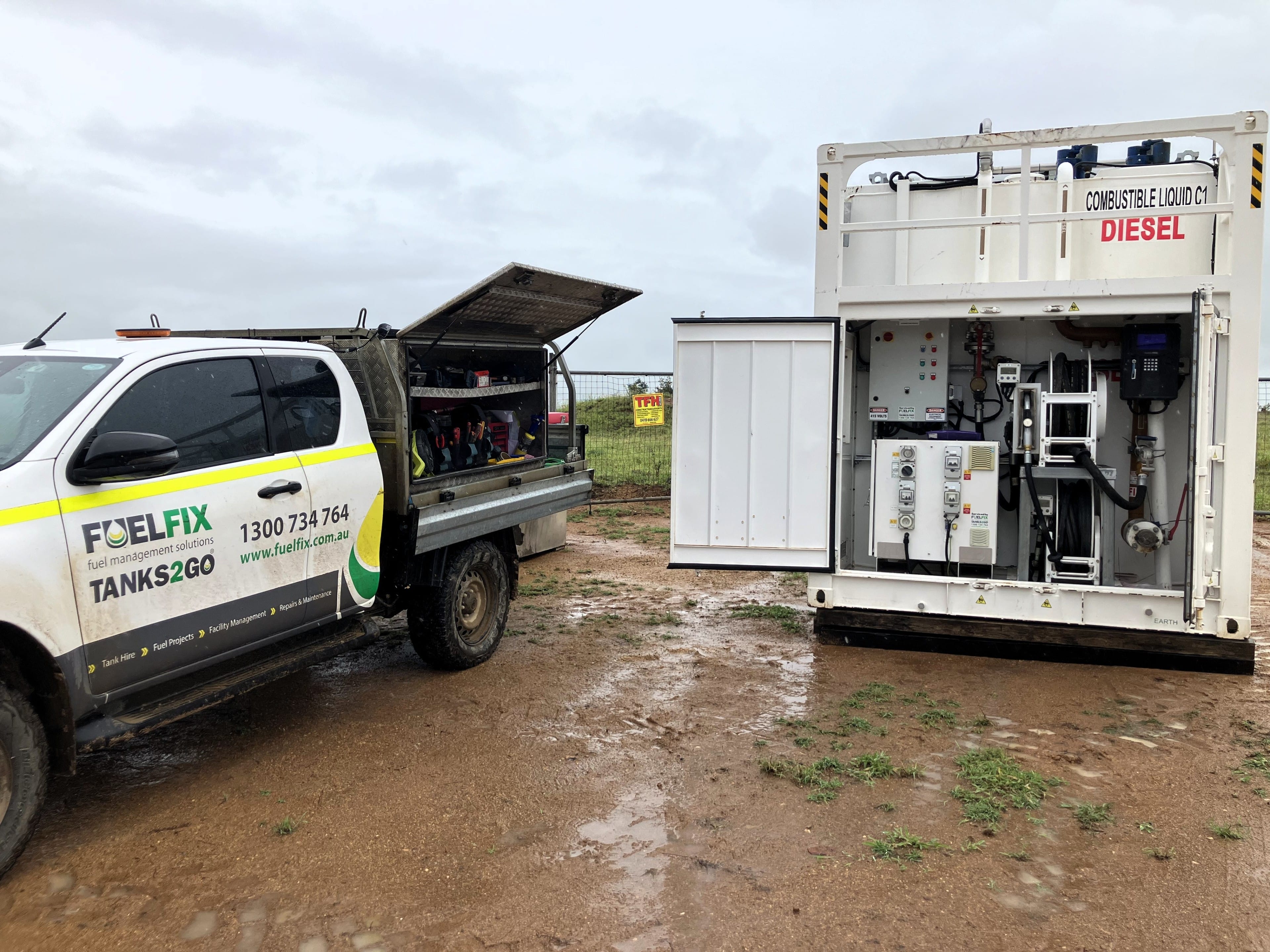 A Fuelfix service vehicle and a solar-powered fuel tank system operating on a remote, muddy construction site. The tank features solar panels and battery energy storage system, replacing diesel generators to power fuel dispensing for service vehicles, reducing emissions and operational costs. the door is open, and you can see the pump system powered by the battery.